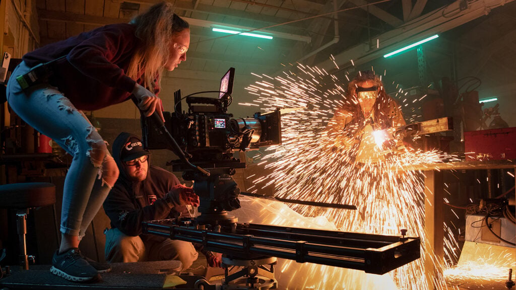 Woman films a welder with camera on rails while a man looks on