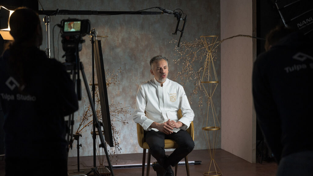 A man in a white chef's jacket sits on a chair in a studio while a camera films him