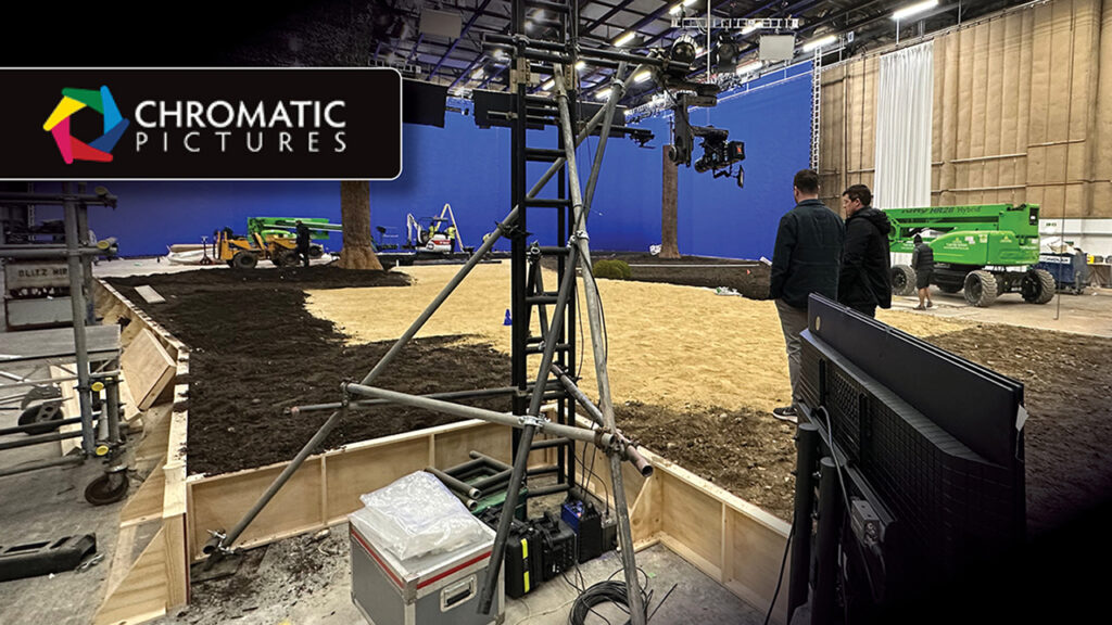 two men stand on a film set with dirt, trees and a bluescreen
