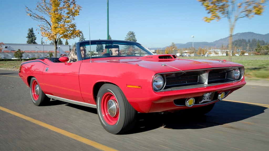 A man and a woman ride down the street in a red droptop Cuda muscle car
