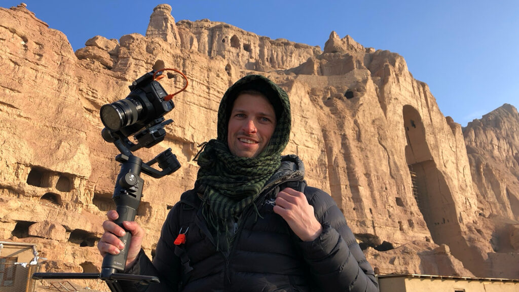 a man stands holding a Blackmagic Pocket Cinema Camera on a tripod in the mountains of Afghanistan