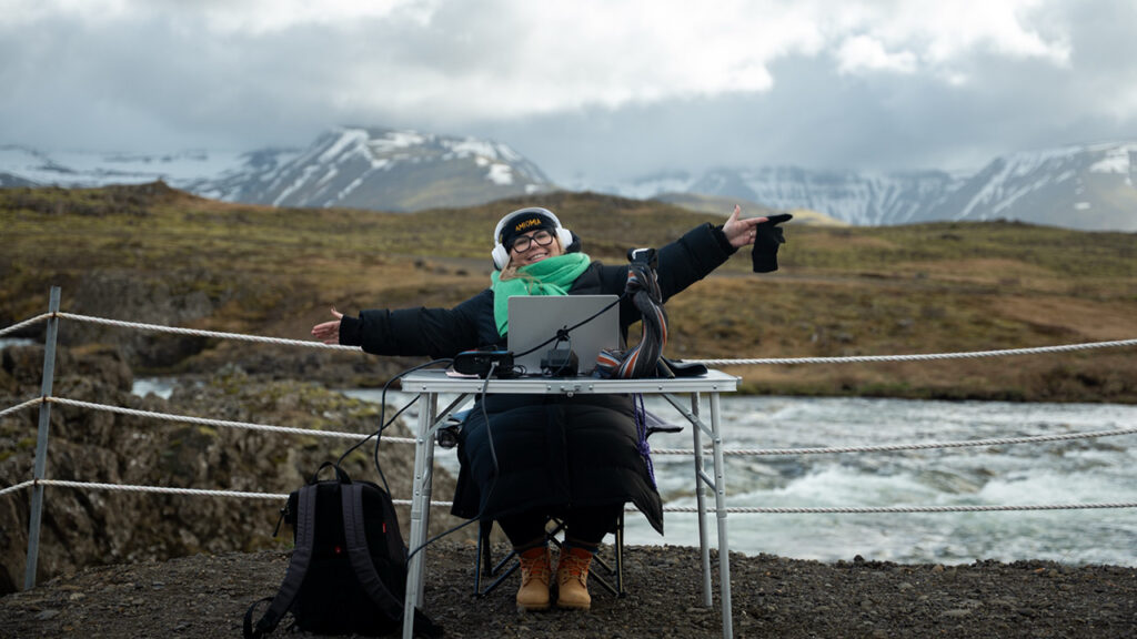 A woman sits at a desk with a laptop and production equipment in the outdoors of iceland