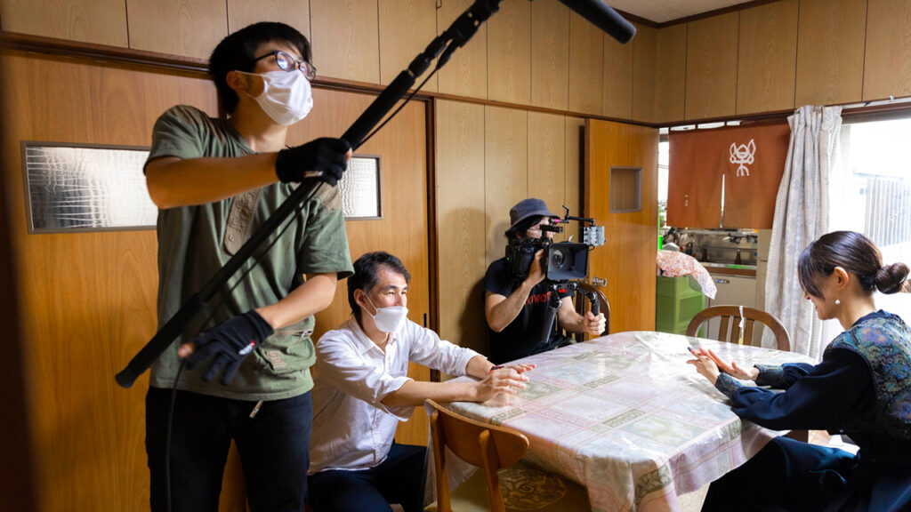 a person holds a boom while a man operates a camera to film a woman sitting at a table