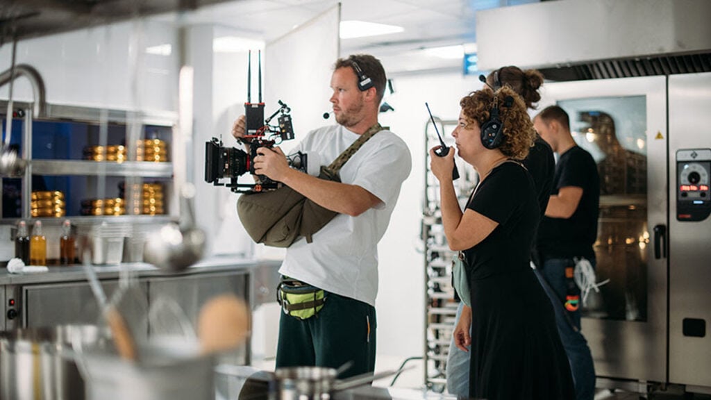 a man operates a camera while a woman speaks into a walkie talkie in a industrial kitchen