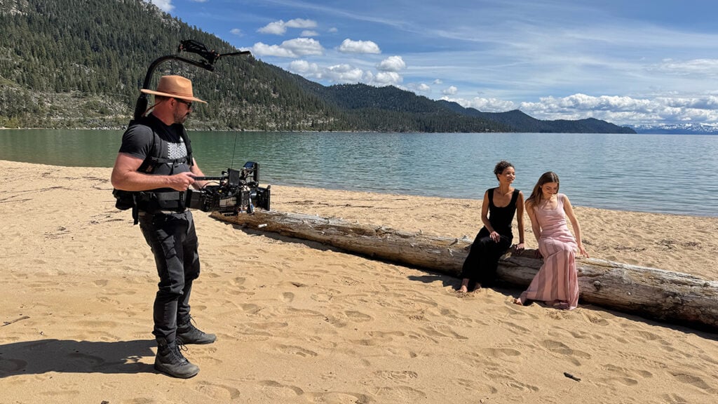 A man holds a camera pointing it at two women sitting on a log on the beach