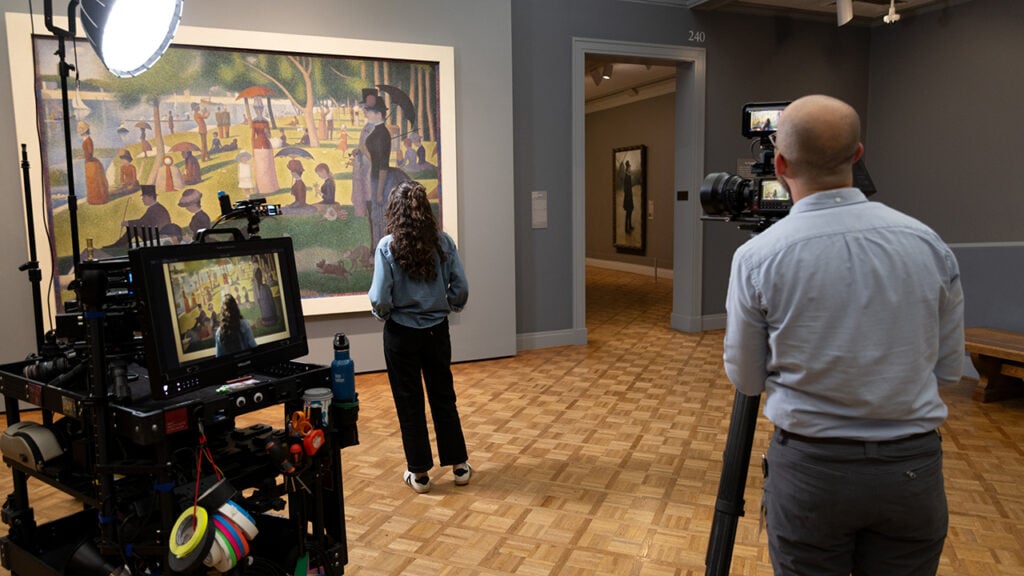 A man films a woman who is looking at a large piece of art
