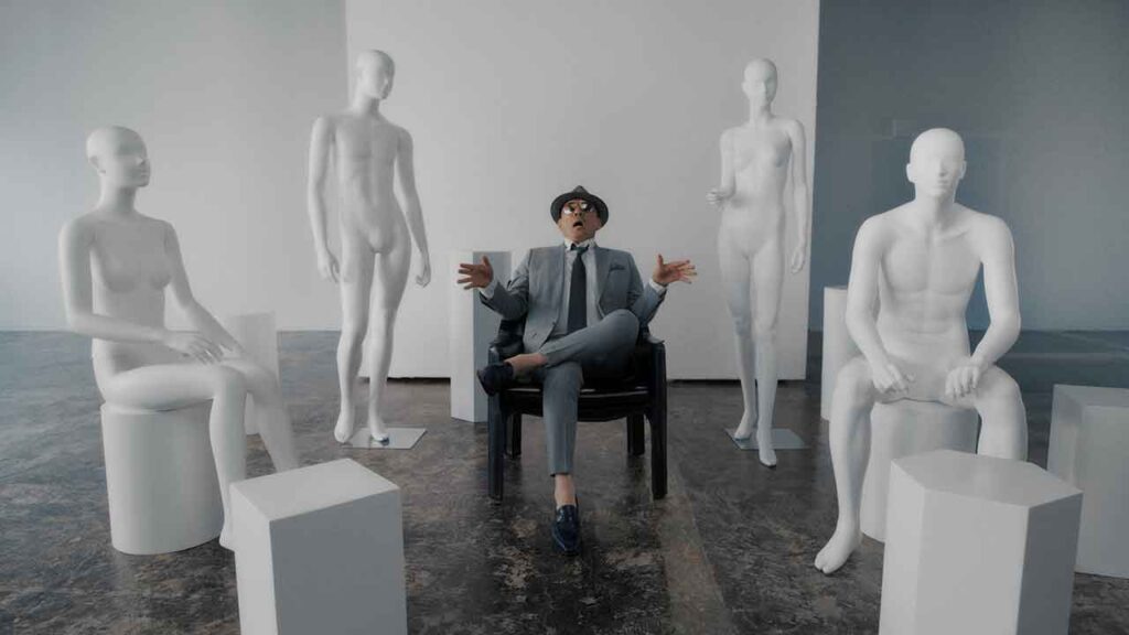 shot from a music video with a man in a grey suit sitting in a room full of mannequins