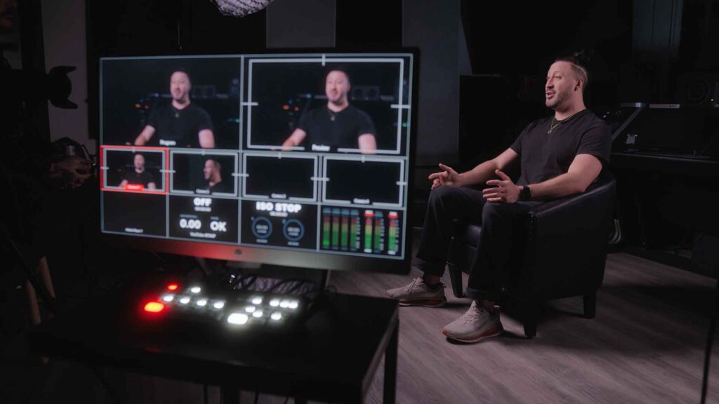 A man sits on set while also appearing on a monitor display