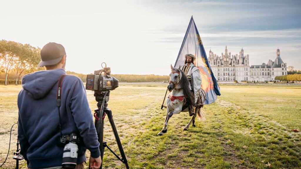 one man films with a Blackmagic camera while another man rides a horse while holding a large flag