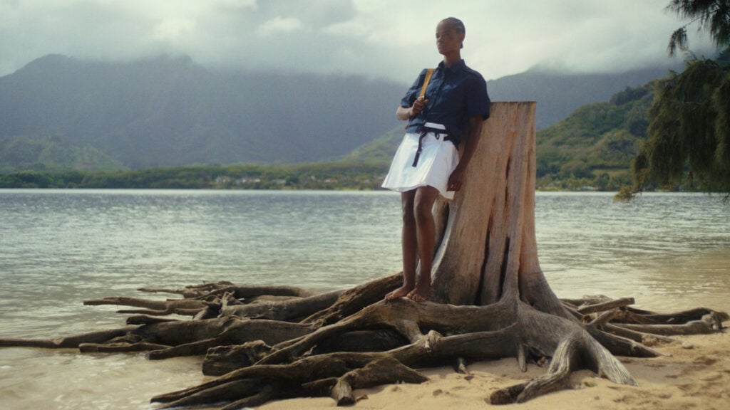 A woman leans against a tree stump next to a body of water