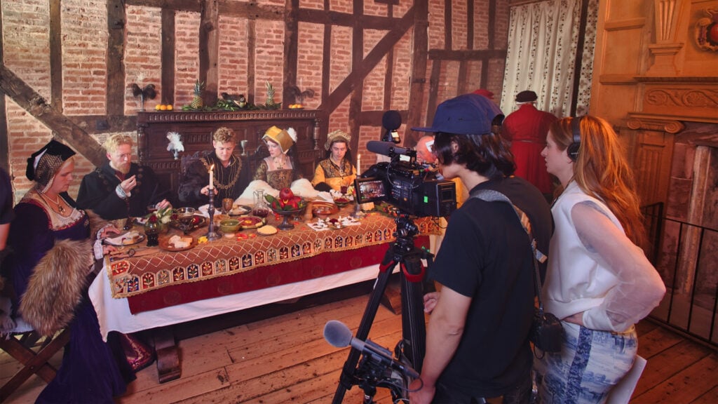 filmmakers stand on a Tudor themed set filming actors eating at a table