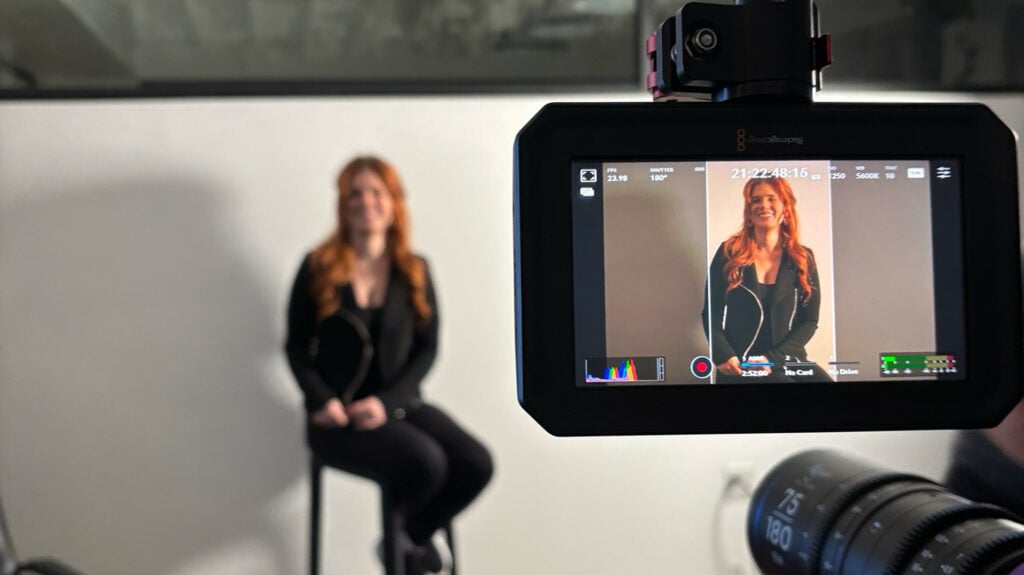 A woman sits on set in a white cove and her image is displayed in a camera monitor
