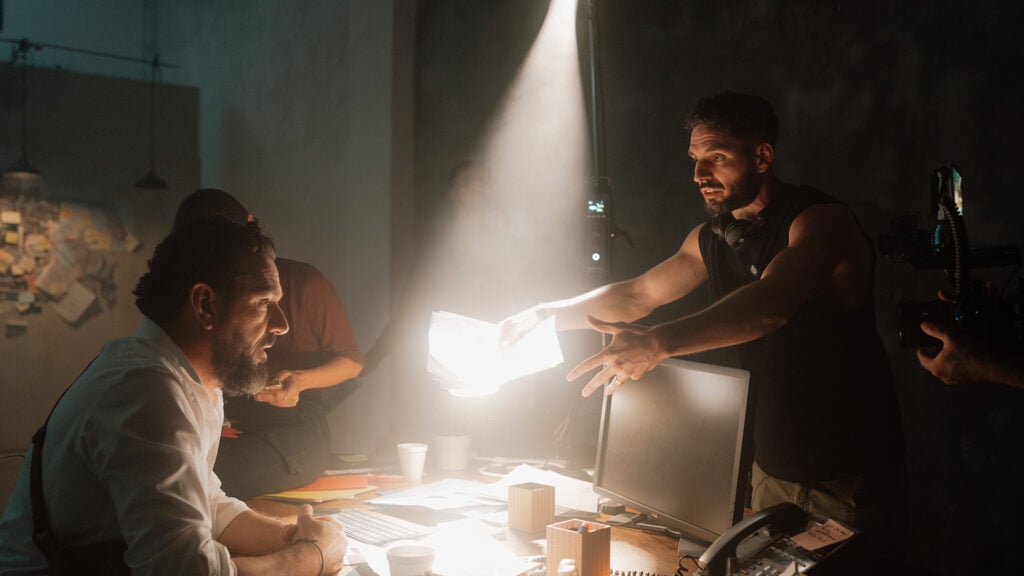 on the set of a movie, the director stands above two people who sit at a table lit from above
