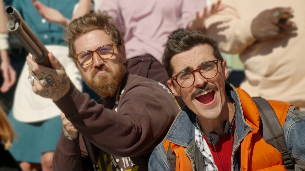two men look at the sky, one holding a shotgun