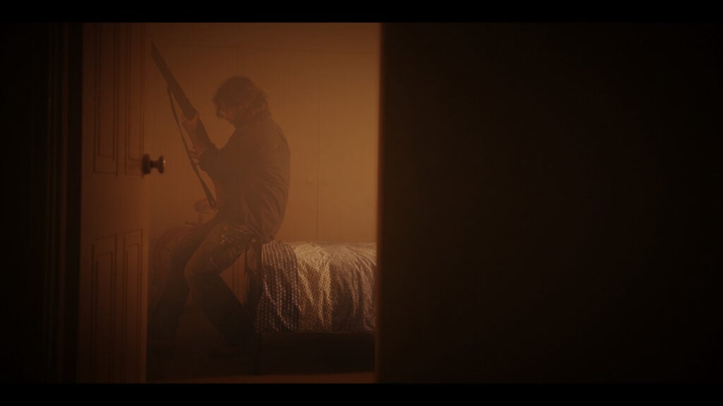 a scene from the film Where Dead Things Grow showing a man sitting on the edge of a bed with a shotgun