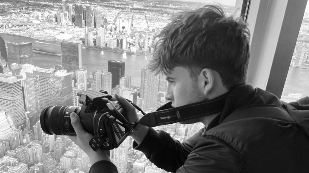 A young man points a camera out the window at a city skyline