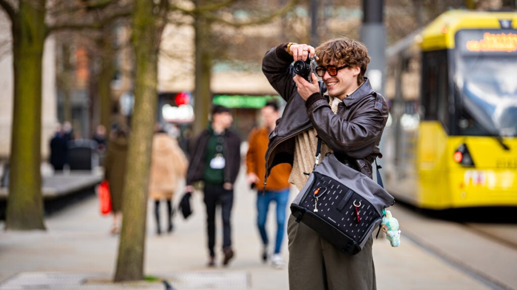 A man uses a camera to take a picture on the sidewalk with a Manfrotto Uncover Messenger bag on his shoulder