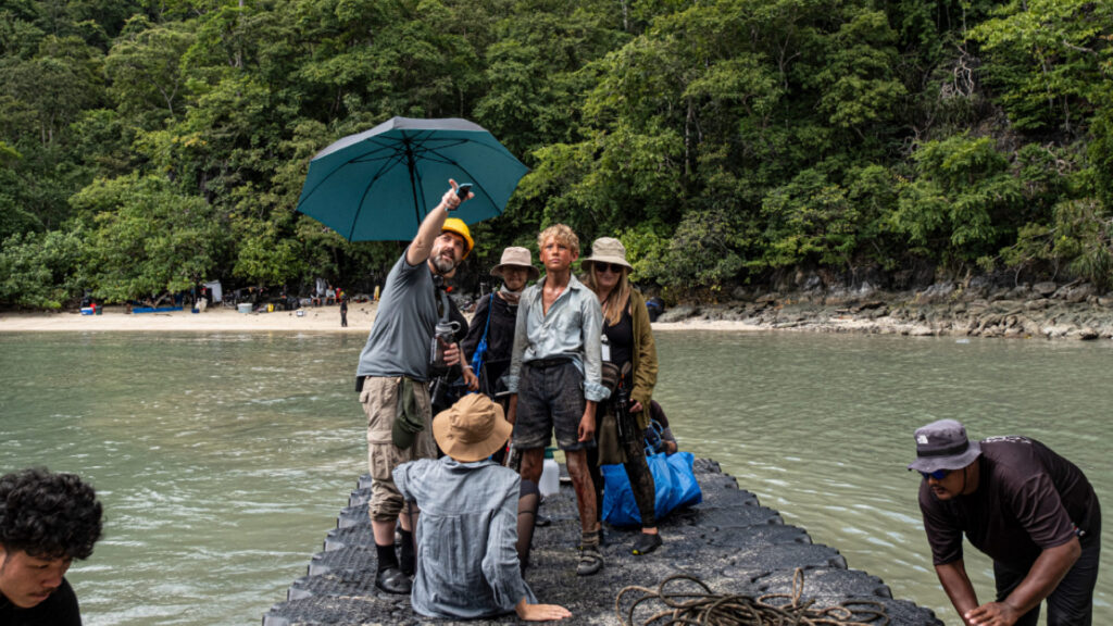 a group of actors and production crew stand on a raft in a lagoon in the jungle