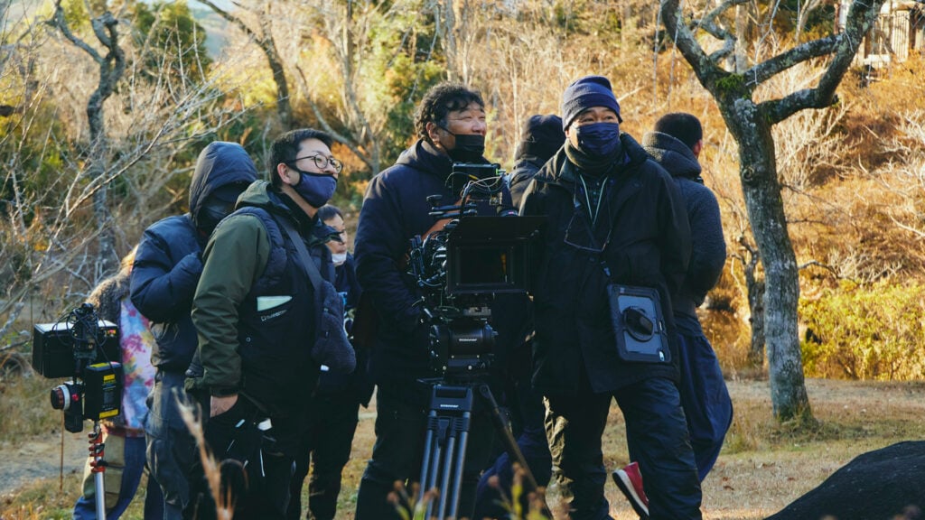 a production crew with cameras looks toward the set while standing in a wooded area