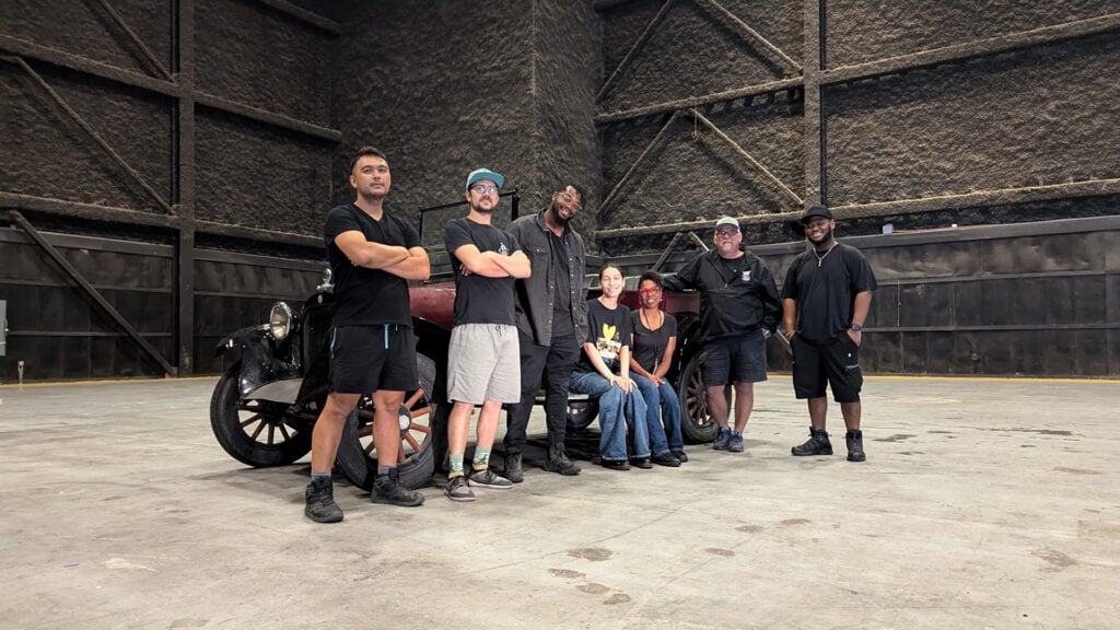 The BTS filmmaking crew stands in front of an old car