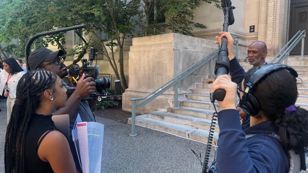 a cameraman operates a Blackmagic camera while another person holds a boom while filming a man talking in front of a large set of stairs